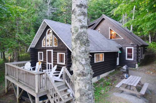 A cozy wooden cabin surrounded by trees, featuring a deck with chairs and a picnic table in the yard.